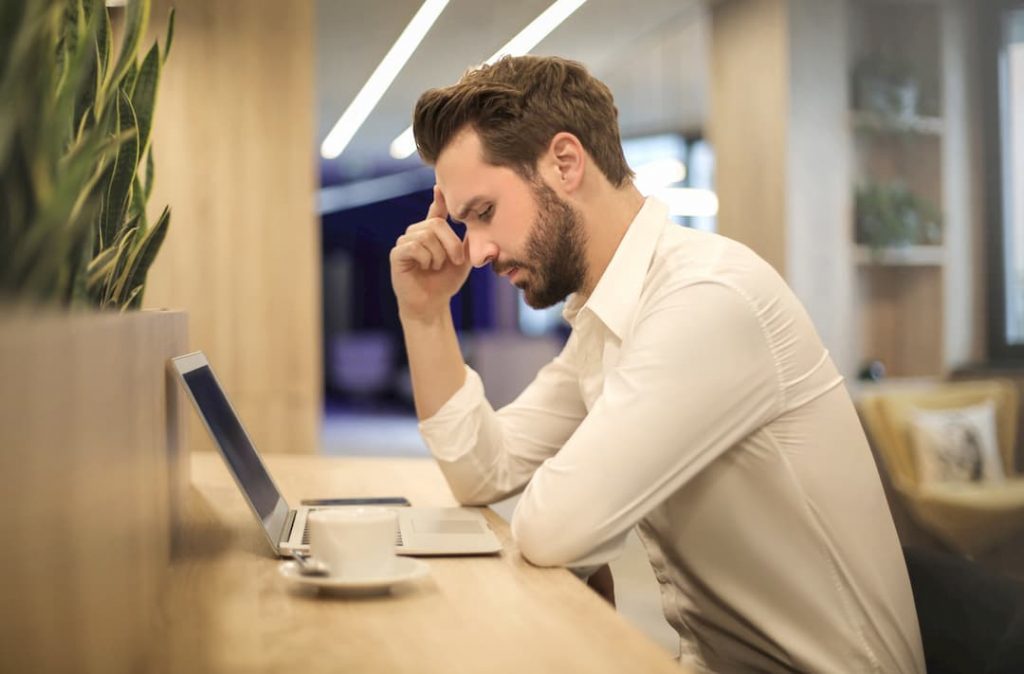 Young man sitting at desk with laptop and coffee looking stressed, office workers risk migraines sickness at work, e-Surgery