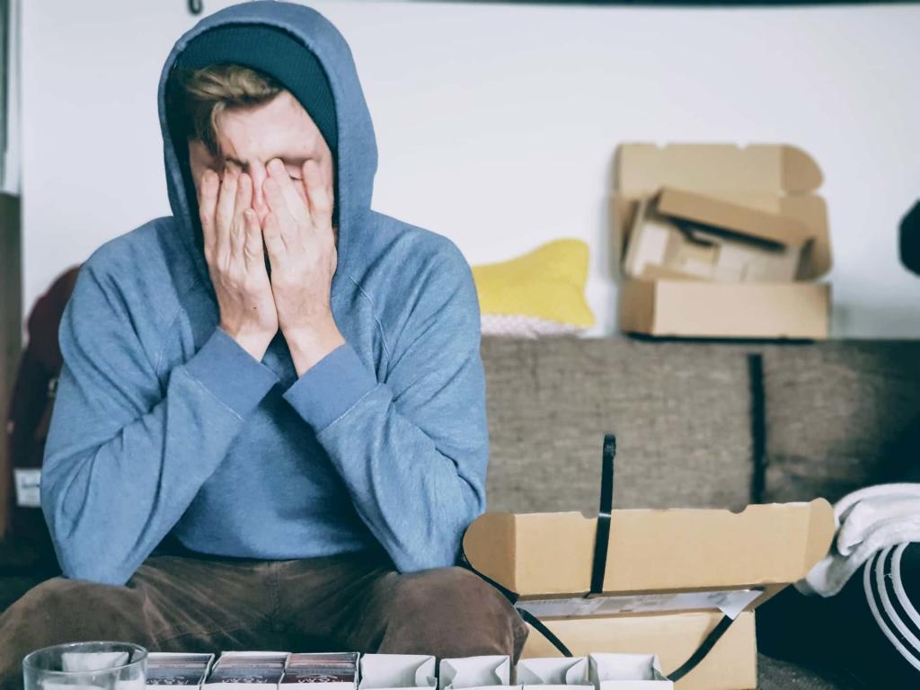 Young male wearing a hoody sitting on a sofa with brown cardboard boxes around him. He has both of his hands covering his face and looks sad | More Young People Than Ever Will Be Lonely This Christmas | e-Surgery 