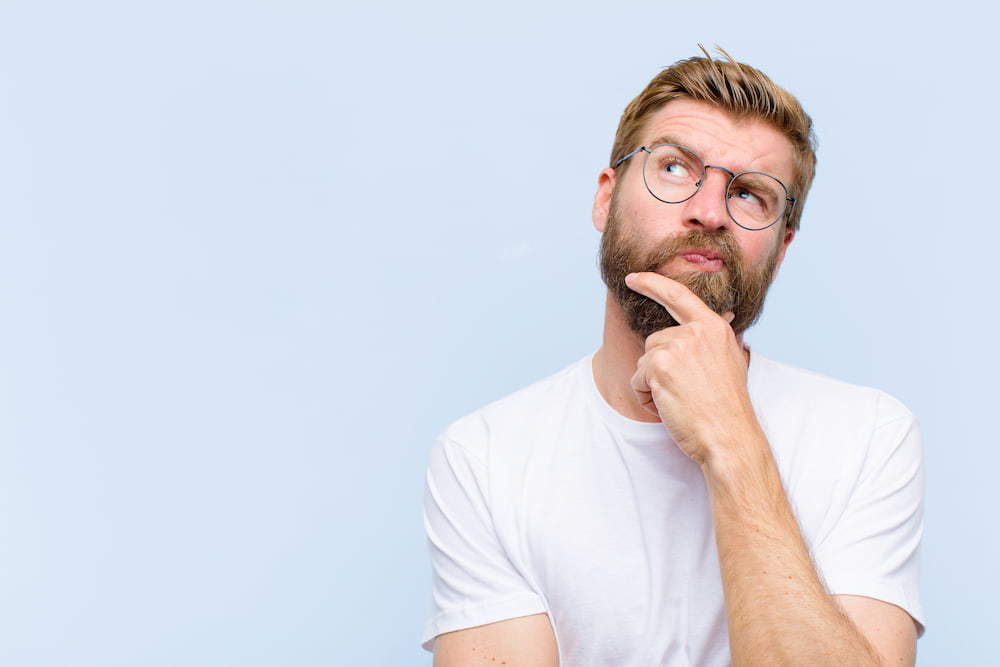 Young male with a bears wearing glasses and a white t-shirt looking interested. He has his chin rested in his thumb and index finger | The Male Pill: Myth Or The Future Of Contraception? | e-Surgery 