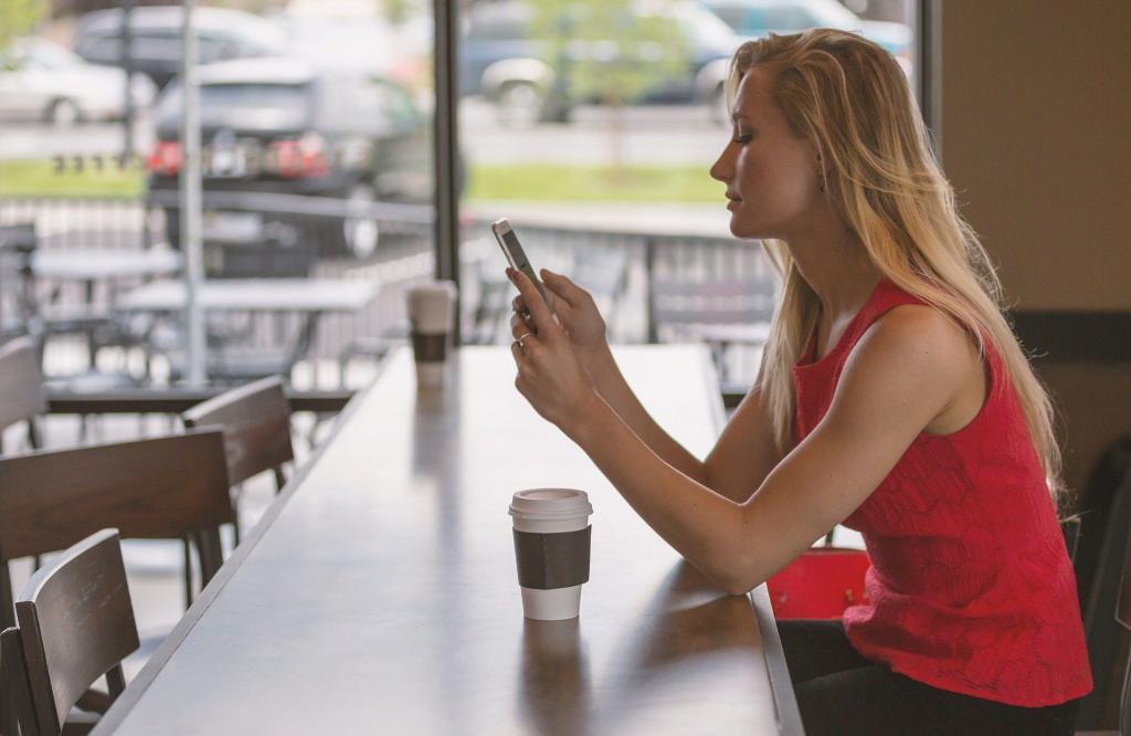 Young blonde female in a red vest top sitting in a coffee shop on her phone | The (almost) A-Z of Contraception | e-Surgery 