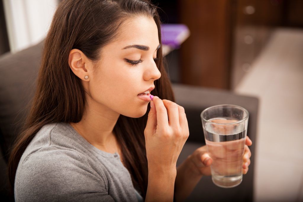 Young female with long brown hair sitting on a sofa putting a pill in her mouth. She is holding a glass of water | The Truth About The Morning After | e-Surgery 