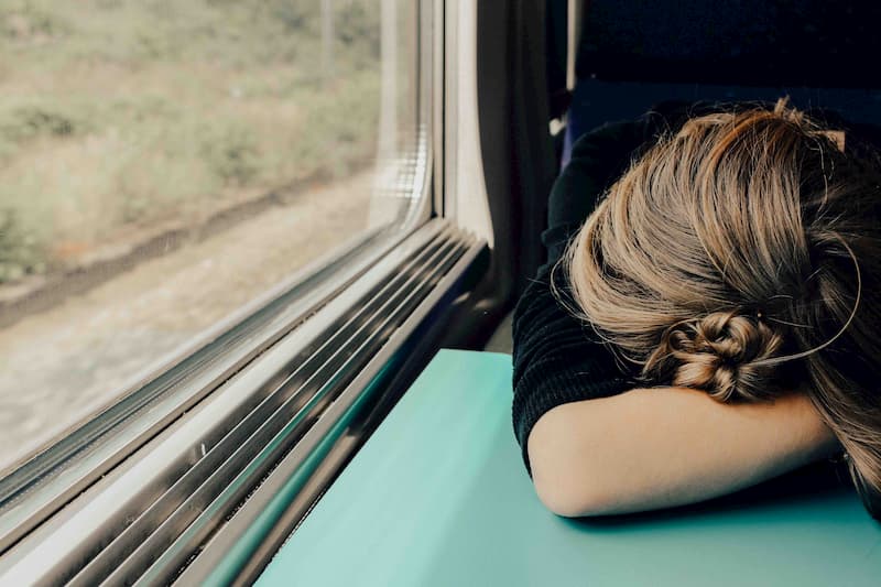 Blonde woman on a train resting her head on the table and covering her face | Tired All The Time? 6 Common Anaemia Symptoms | e-Surgery 