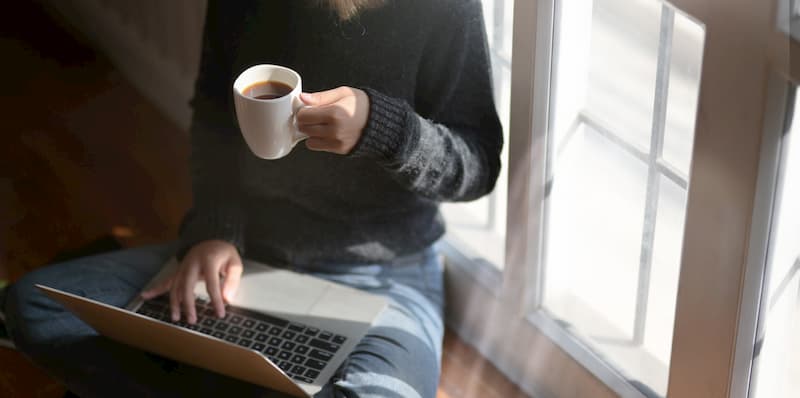 Person drinking coffee working from home on laptop, welling in self isolation, e-Surgery