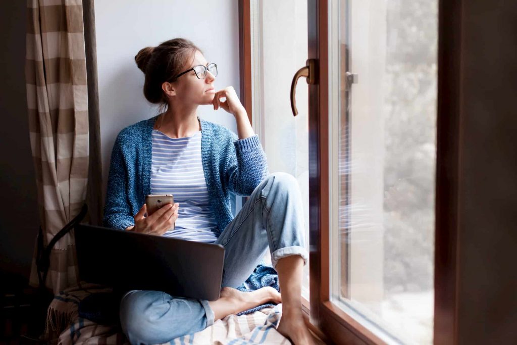 Woman looking out of window holding phone and laptop, ordering medication online in lockdown, e-Surgery