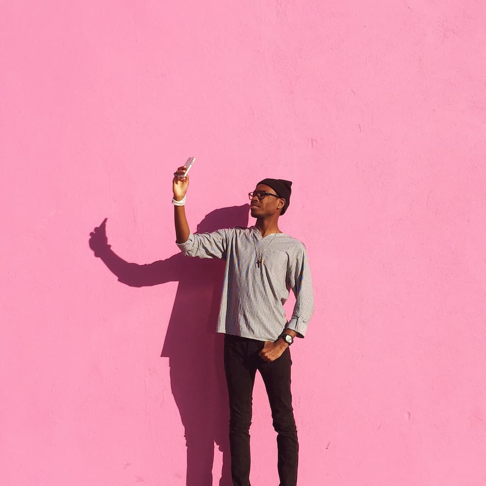 Young man in beanie hat against pink background holding up mobile phone to take selfie, social media in lockdown, e-Surgery