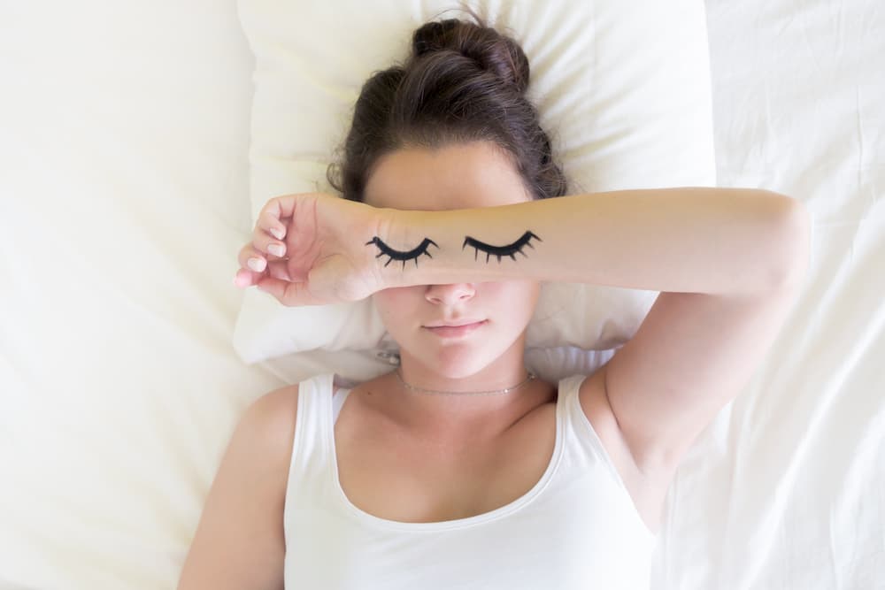 Woman with arm over face in bed and closed eyes with eyelashes drawn on, sleep is important to help acne in lockdown, e-surgery