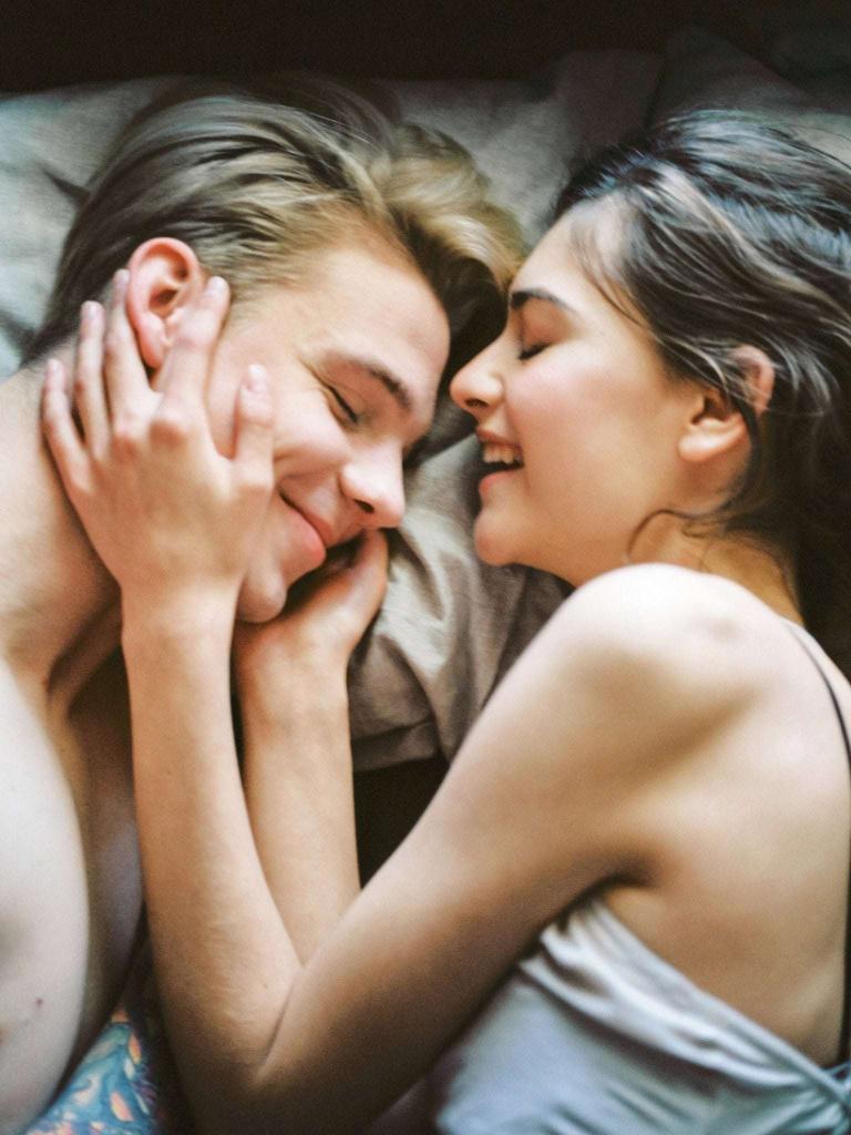 Young couple laying in bed, female has her hands either side of the males face. Both individuals are smiling and look happy | Why Do Young People Keep Their STIs a Secret? | e-Surgery