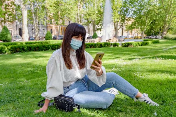 Young student, woman with brown hair, sitting on grass with phone in hand and a mask, guide to student health, e-Surgery