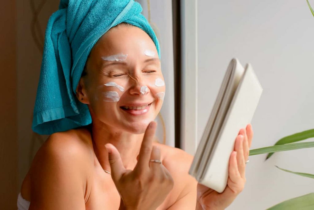 Female smiling with eyes shut wearing a towel on her head applying face cream. 