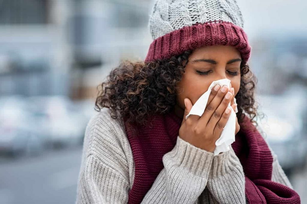 Young black woman wearing a knitted hat and scarf sneezing into a tissue | How To Deal With Winter Asthma | e-Surgery 