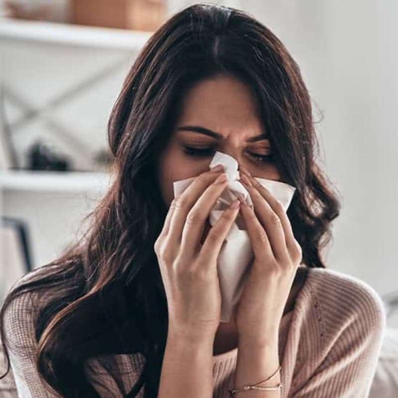 Mild hay Fever: Symptoms & Treatments, young woman sat on a sofa sneezing with a tissue covering her face | e-Surgery