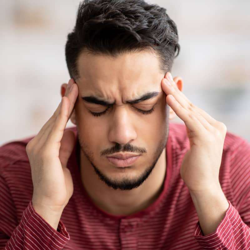 Asian man holding the bridge of his nose with one hand and his glasses with another. He is and scrunching his face up in pain | Does Your Diet Trigger Migraines? | e-Surgery