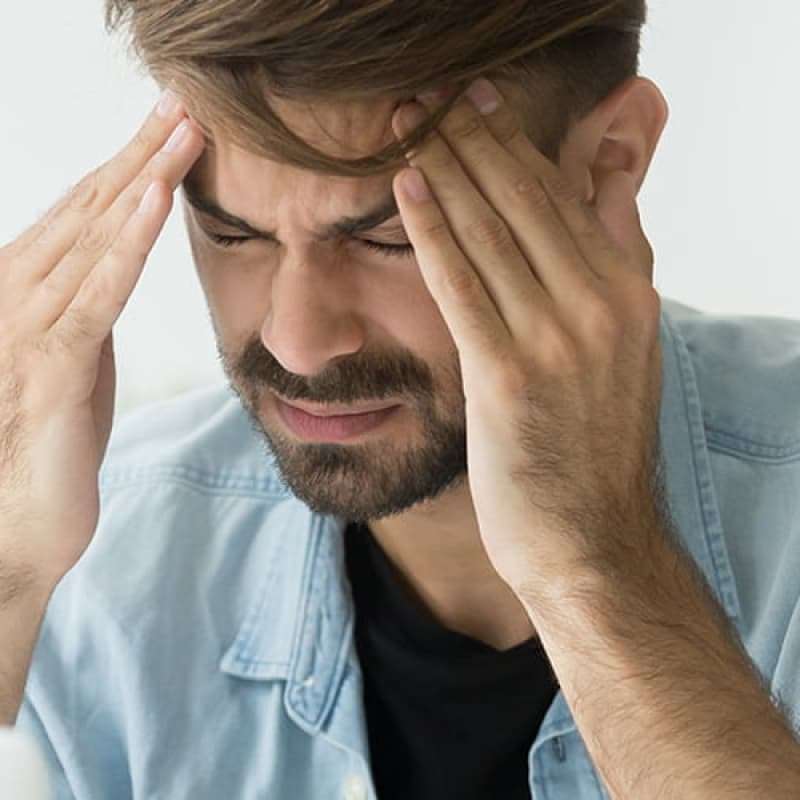 Home remedies for migraines, middle aged bearded man sitting at his laptop with his fingers pressed against his temple looking stressed | e-Surgery