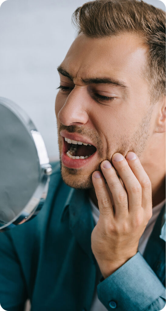 A man looking at his teeth through the mirror and holding his jaw looking uncomfortable